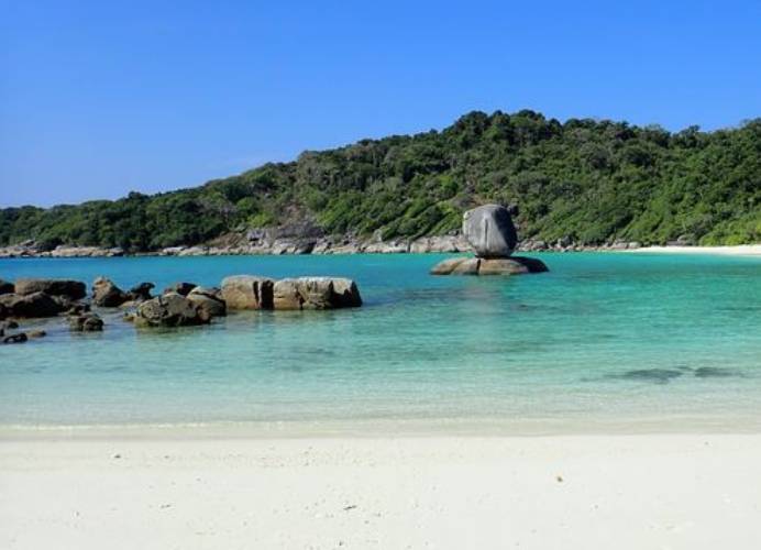 Playa desierta de arena blanca y agua turquesa. Hay algunas rocas en el agua y al otro lado de la bahía se ve la selva.